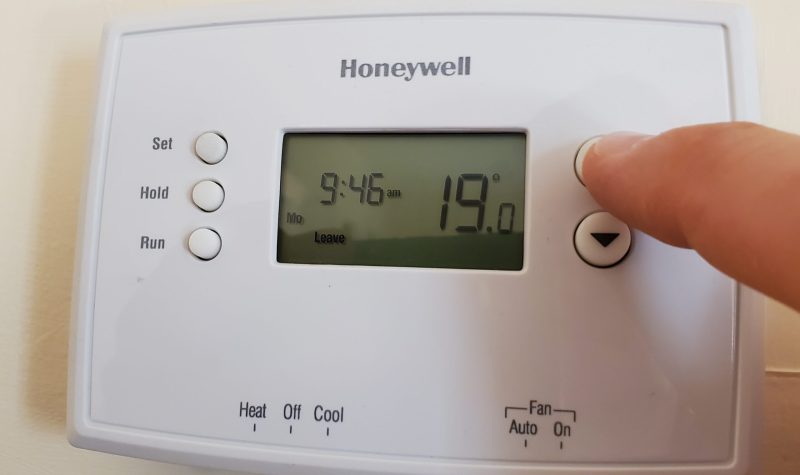 A white Honeywell thermostat on the wall with a finger pressing a button.
