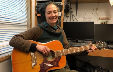 A smiling woman in an earth tone sweater sits with a burnt orange acoustic guitar.