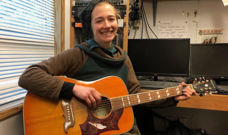 A smiling woman in an earth tone sweater sits with a burnt orange acoustic guitar.