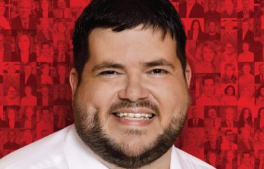 Homme cheveux brun foncé, courte barbe souriant avec chemise blanche sur fond rouge