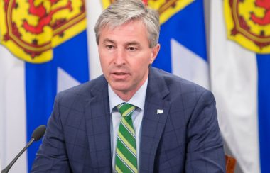 A man sits at a desk in front of a row of Nova Scotia flags