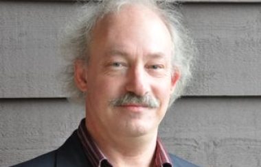 A head shot of a man with grey hair and a moustache wearing a red collared shirt and grey jacket.