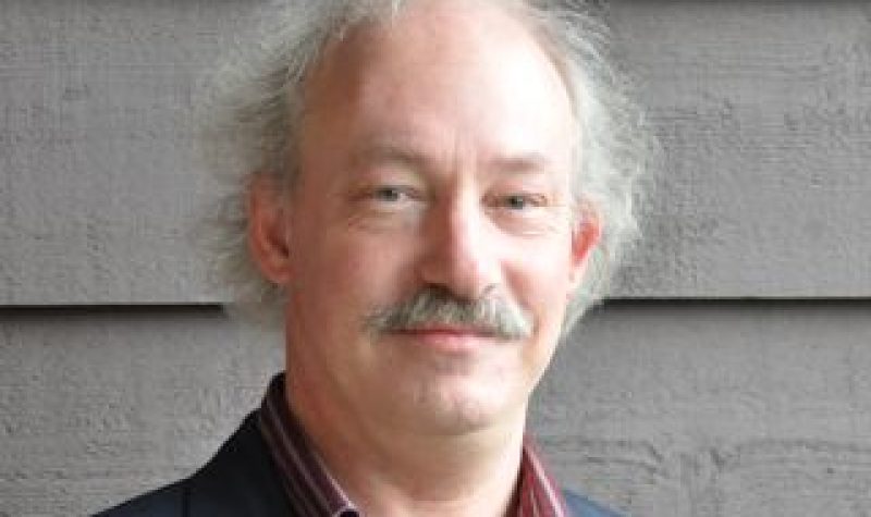 A head shot of a man with grey hair and a moustache wearing a red collared shirt and grey jacket.