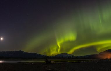 curtains of green and yellow cross the sky at night over a lake near Terrace BC. On the right bottom of the phot is the orange light of a campfire in the distance as well as the moon on the left side of the horizon of a mountain in blackness.