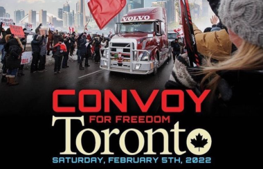 A truck on a road surrounded by a crowd of people holding red and white Canadian flags with buildings in the background.