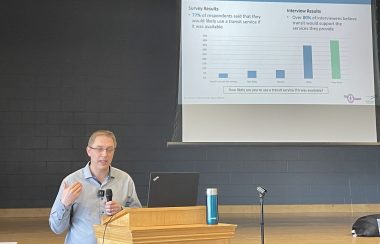 Man at podium in light blue/grey top holds microphone with a large screen behind him showing graph.