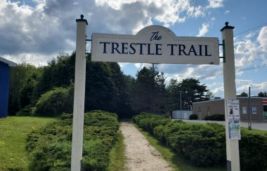 A white and blue sign overhangs the entrance to the Trestle Trail in Liverpool. There are hedges on either side of the gravel trail.