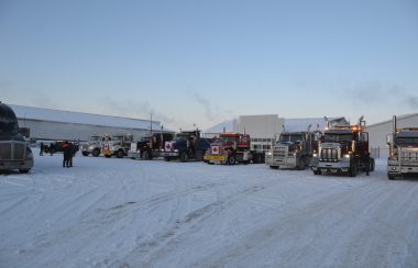 A group of roughly a dozen big rigs idling in a snowy parking lot, some with Canadian flags attached to the grille.