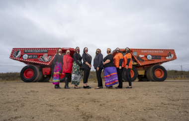 Indigenous women wearing colorful ribbon skirts in front of the two painted haul trucks used in industrial setting.