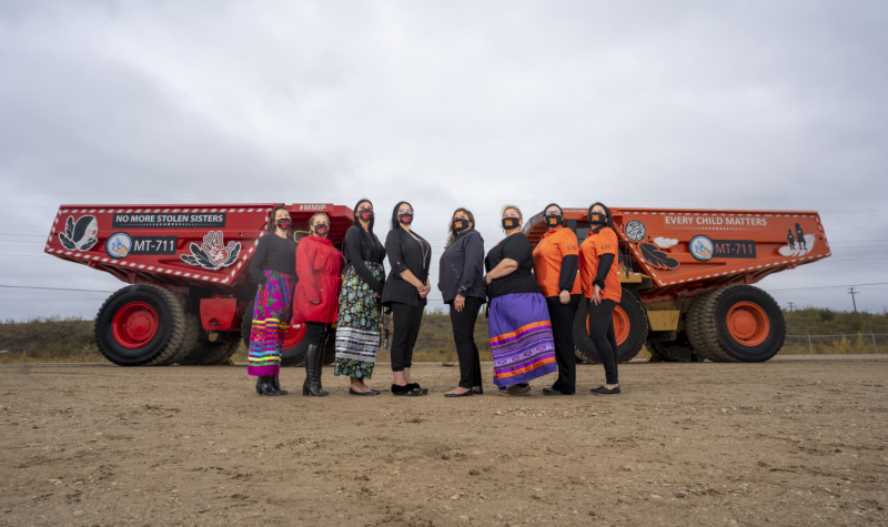 Indigenous women wearing colorful ribbon skirts in front of the two painted haul trucks used in industrial setting.