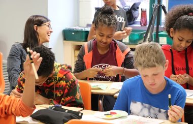 Young children work on bead art with a laughing older woman in a well-lit classroom.