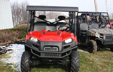 A photo of two large (compact car sized) four wheeled all terrain vehicles on grass and snow. The vehicle in the forground has an orange hood, crash bar, and heavy treaded tires, with an exposed cockpit with a roof that seats two. The vehicle in the background is similar to the other vehicle, but it has a camouflage-like colour scheme on its hood, and its two person cockpit is roofless. More vehicles are parked further afield.
