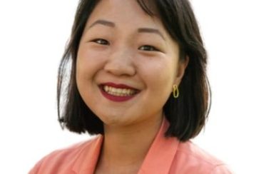 A person with black hair is smiling. They are wearing a pink blazer and standing in front of a white background.