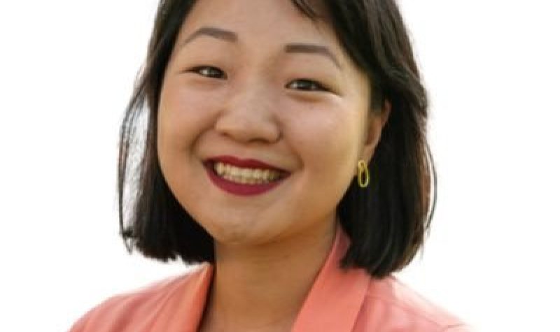 A person with black hair is smiling. They are wearing a pink blazer and standing in front of a white background.