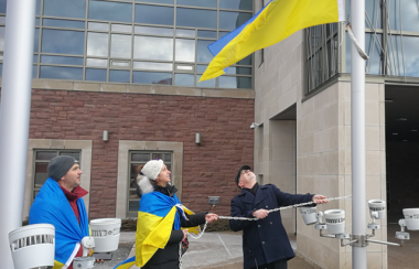 Two people wrapped in blue and yellow flags and one person dressed in black, two of whom are hoisting a blue and yellow flag on a flagpole.