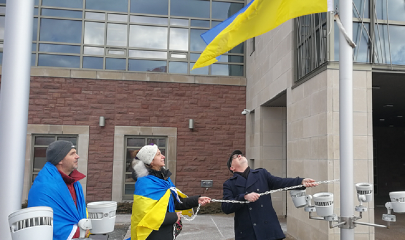 Two people wrapped in blue and yellow flags and one person dressed in black, two of whom are hoisting a blue and yellow flag on a flagpole.