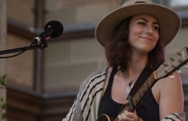 Une jeune femme portant un chapeau et un chandail brun jouant de la guitare en avant d'un micro.