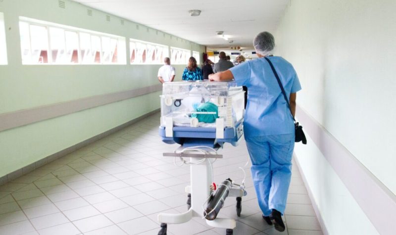 A healthcare worker transports a patient down a hallway wearing scrubs.