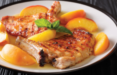Fresh peaches sit on a barbecued pork chop with a greenery garnish on a white plate.