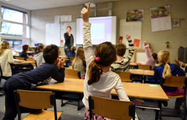 Young students raise hands in a daylight-lit classroom.