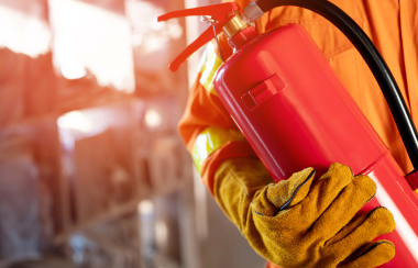 A firefighter holds an extinguisher, red in colour, while wearing thick yellow gloves and orang clothes.