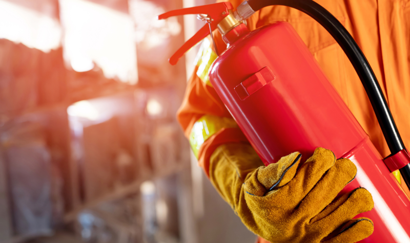 A firefighter holds an extinguisher, red in colour, while wearing thick yellow gloves and orang clothes.