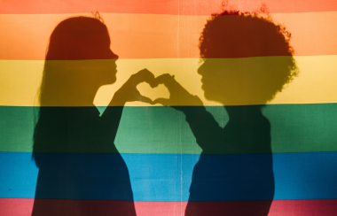 Two children create the outline of a heart with their hands through a rainbow pride flag with a light shone on it.