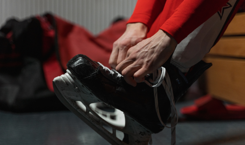 A hockey player dressed in red ties up a skate in a dim-lit dressing room. A bag of equipment sits in the background.