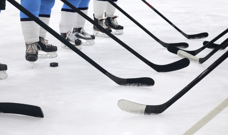 Hockey players photographed from the knees down stand idle on the ice with their sticks surrounding a single puck.