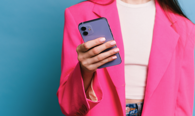 A woman in front of a blue background in a pink blazer holds a cell phone.