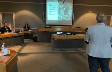 CEO of CMHA Waterloo Wellington Helen Fishburn stands before Wellington County council with a presentation on a screen in a dim-lit council chamber.