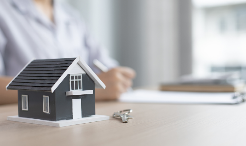 A model house sits with a pair of keys. In the background a person fills out what appears to be a lease.