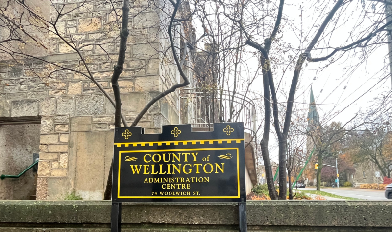 The County of Wellington Administration Centre sign in black and yellow stands before a brick building and trees on a fall day.
