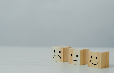Three wooden blocks have contrasting happy, straight and sad faces on the front of them in front of a blank background.