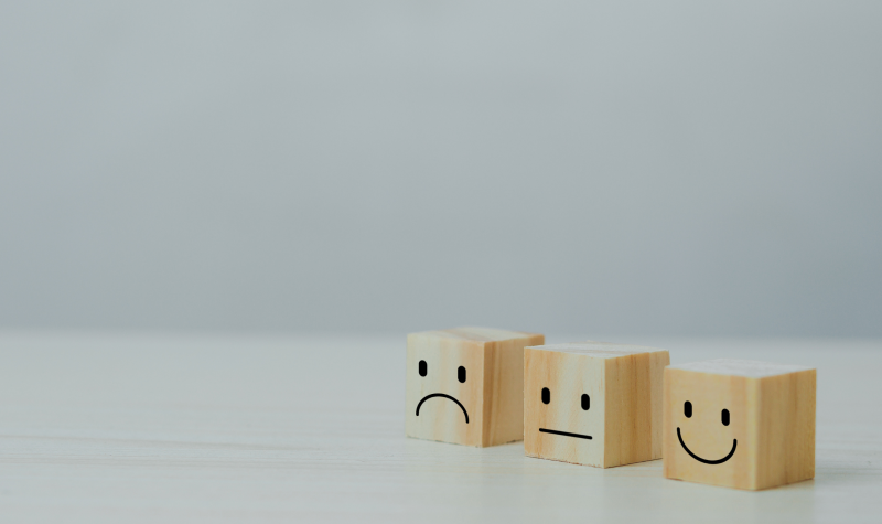 Three wooden blocks have contrasting happy, straight and sad faces on the front of them in front of a blank background.