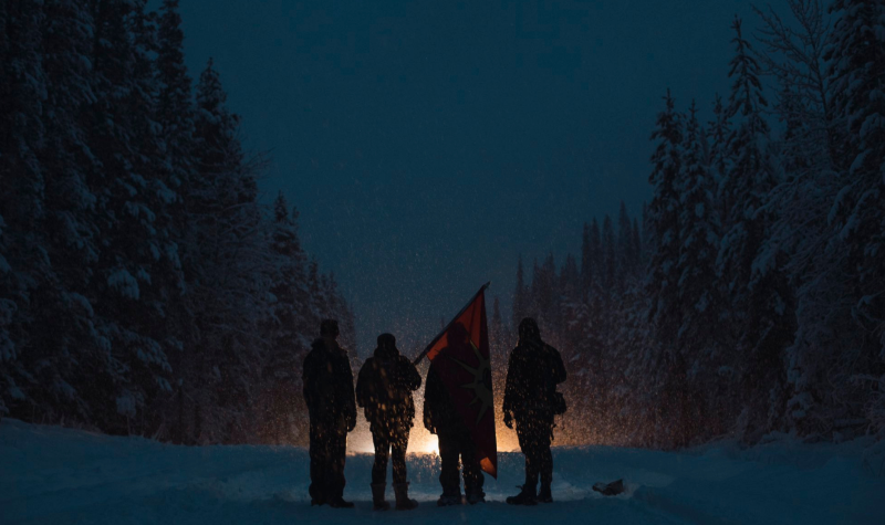 A group of people in winter attire stand before a light at a crossroads in a winter scene with an orange flag.