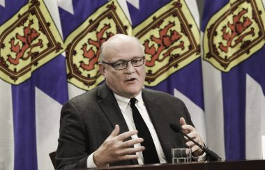 A man sits at a desk in front of a row of Nova Scotia flags