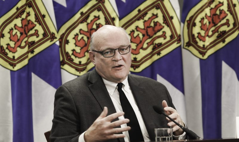 A man sits at a desk in front of a row of Nova Scotia flags