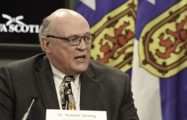 Dr. Robert Strang stands at a podium during a press conference with Nova Scotia flags behind him