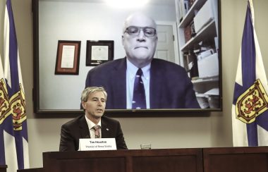 A man sits at a podium while behind him, another man appears on a large screen