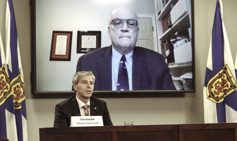 A man sits at a podium while behind him, another man appears on a large screen