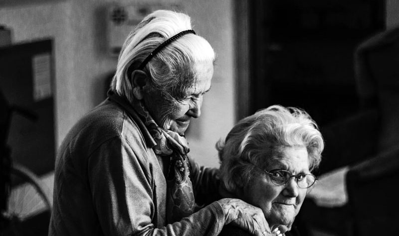 A black and white photo of two seniors. One stands behind the other with their hands on their shoulders as they sit.