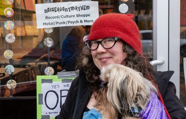 A woman wearing glasses and a red toque holding a dog in front of a record store.