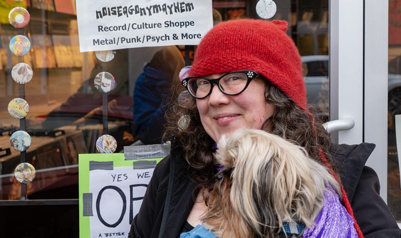 A woman wearing glasses and a red toque holding a dog in front of a record store.