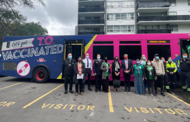 A lineup of people with medical masks on standing in front of a transit bus in a parking lot.