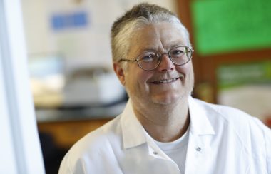 A woman in a white lab coat, smiling, with a blurred background.