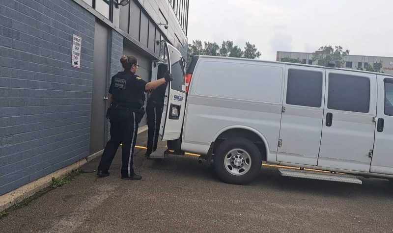 Deux agents policiers en uniforme noir referme la porte arrière du camionnette blanche, lors du journée chaude et nuageuse