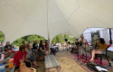 A group of people listen to a woman play a guitar and sing under a large tent.