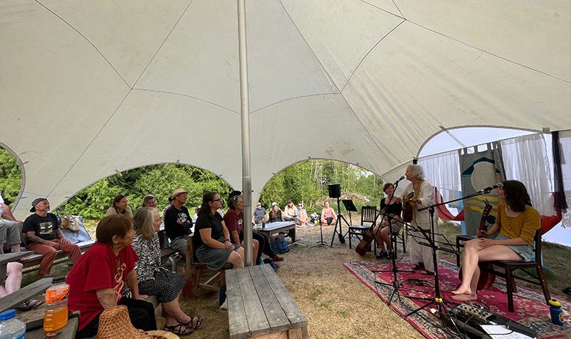 A group of people listen to a woman play a guitar and sing under a large tent.