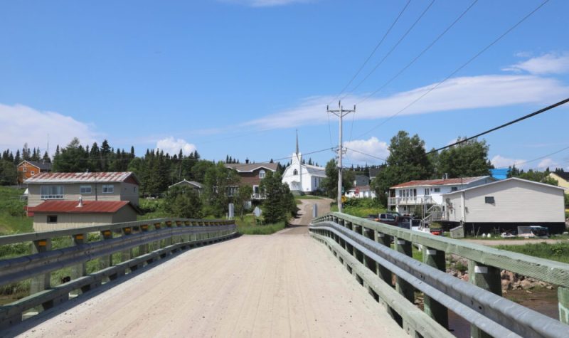 Au cœur d’un village, un pont mène à une église blanche. Quelques maisons modestes reposent autour.